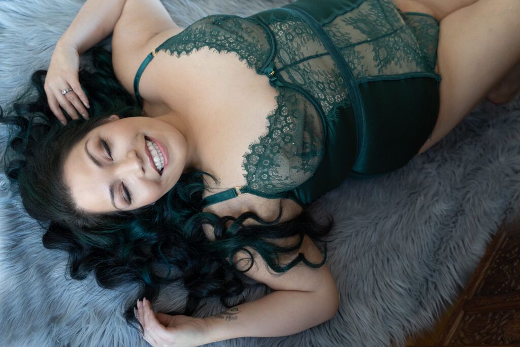 A woman with teal-tinted hair smiles joyfully, lying on a grey fur rug in a lacy green boudoir outfit, showcasing confidence and beauty in a relaxed pose.