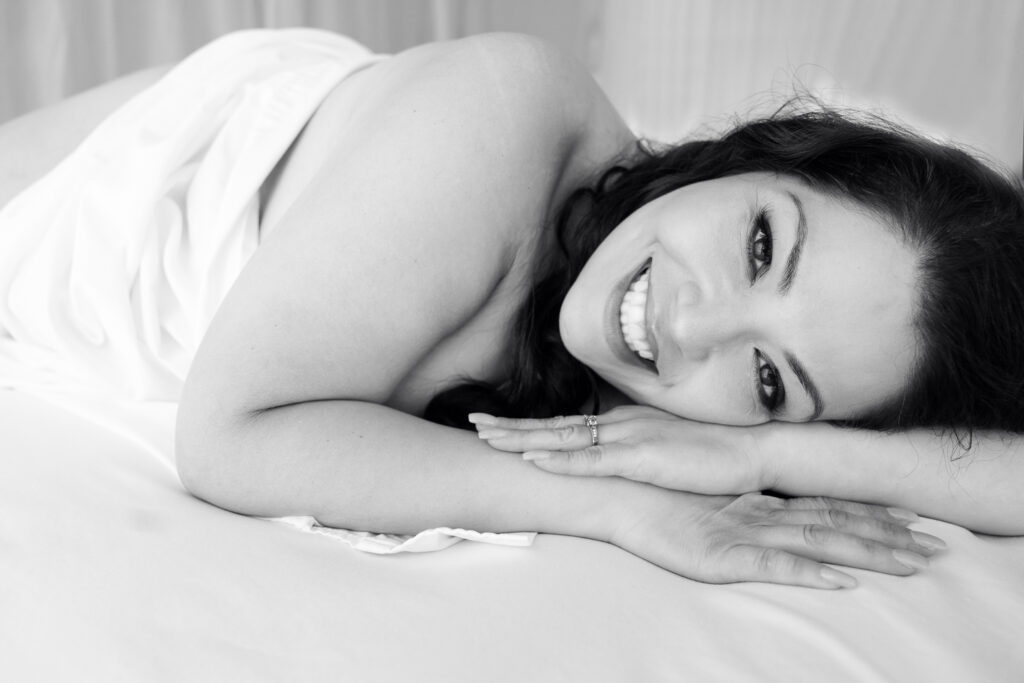 A black and white photo. A woman lays down, smiling widely at the camera, a white sheet covers her chest.